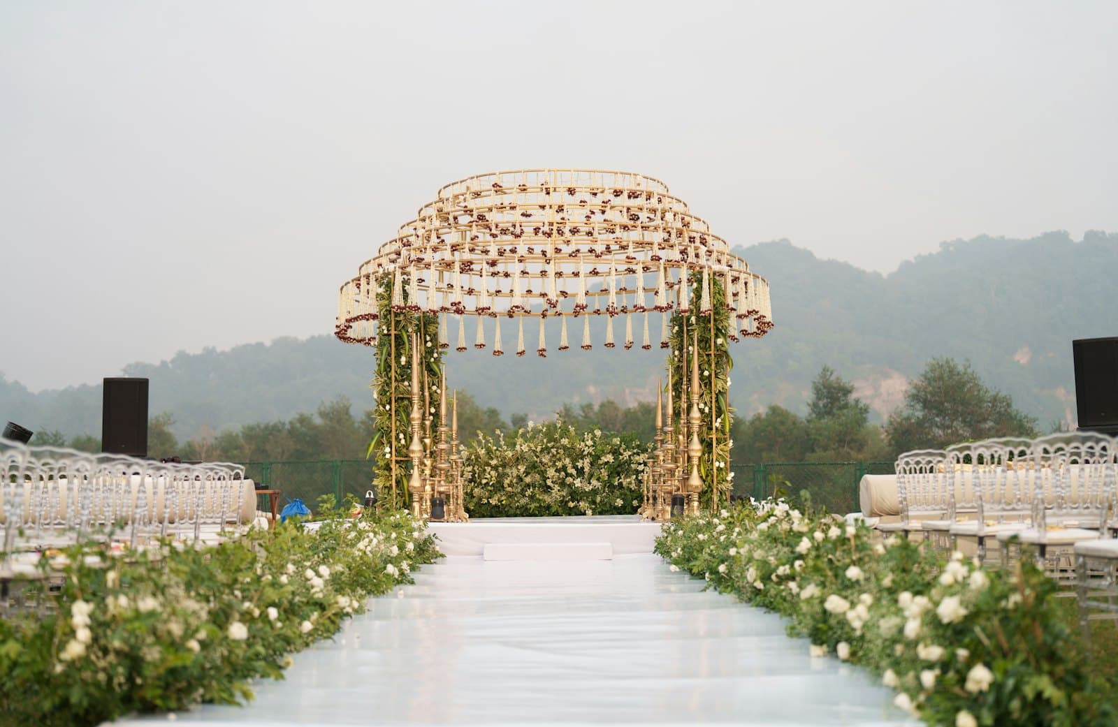 A gold mandap hung with marigold and jasmine strands at the head of a white aisle, chairs set in rows on either side, hills behind.